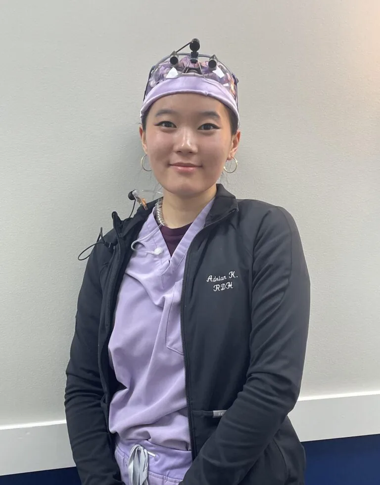 Person wearing a patterned surgical cap and a black jacket embroidered with “Adrian K., RDH” stands in front of a light-colored wall. The jacket is worn over lavender scrubs, and the individual has headgear with attached equipment, suggesting a clinical or dental setting.