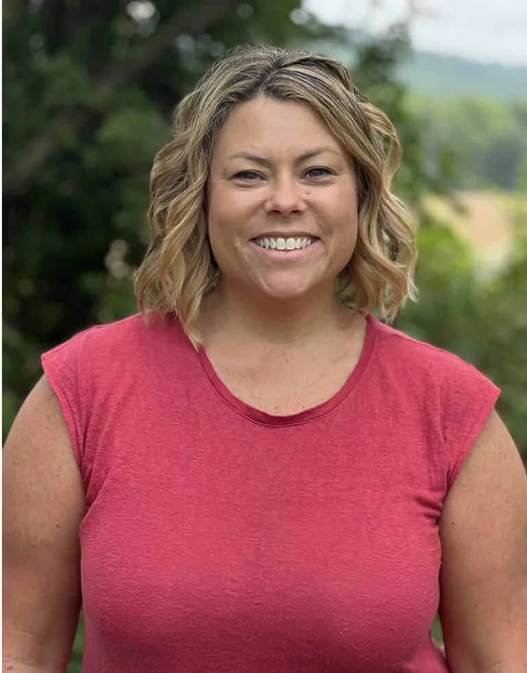 Rachel a receptionist, in a red short-sleeved shirt stands outdoors with trees and distant hills in the background. Their curly hair is visible, and the scene suggests a sunny day in a natural setting.