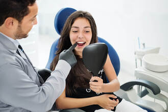 A dental professional wearing gloves examines a patient's teeth during a consultation, with the patient holding a mirror to view their smile. The discussion appears to focus on dental implants as a treatment option for missing teeth.